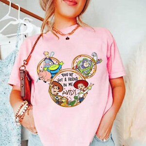 May include: A light pink t-shirt featuring a graphic print of Toy Story characters arranged in a Mickey Mouse ear shape. The center circle contains the text "YOU VE GOT A FRIEND IN ME ANDY" surrounded by illustrations of Woody, Jessie, Buzz Lightyear, Bo Peep, Rex, Slinky Dog, Hamm, and an Alien.