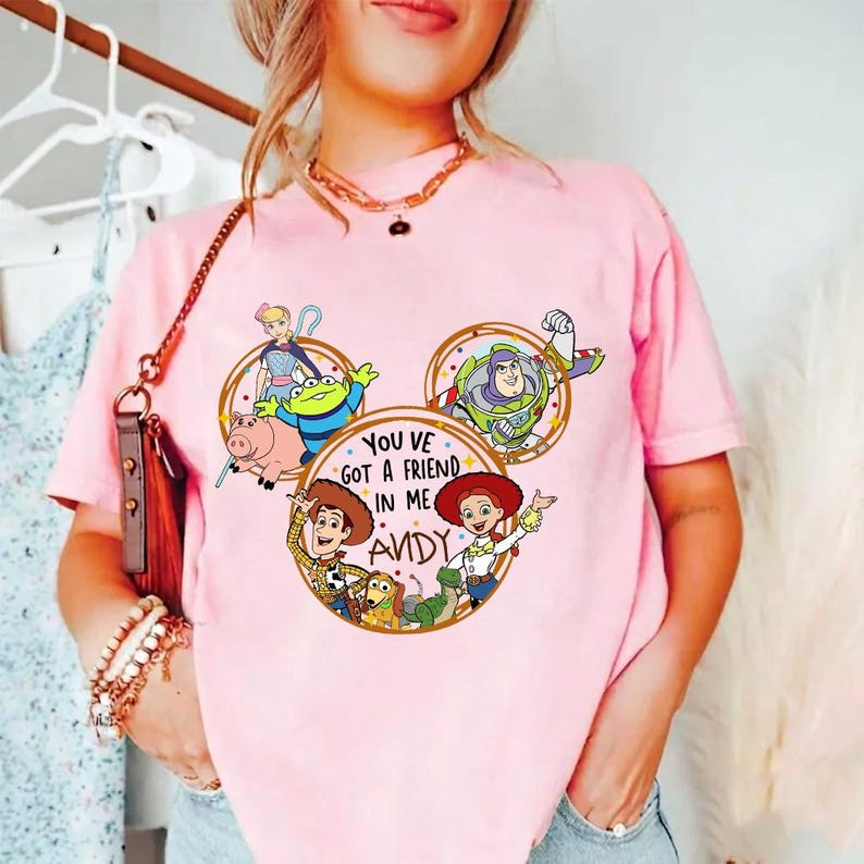 May include: A light pink t-shirt featuring a graphic print of Toy Story characters arranged in a Mickey Mouse ear shape. The center circle contains the text "YOU VE GOT A FRIEND IN ME ANDY" surrounded by illustrations of Woody, Jessie, Buzz Lightyear, Bo Peep, Rex, Slinky Dog, Hamm, and an Alien.