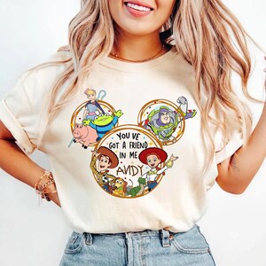 May include: A cream-colored t-shirt featuring a graphic of Toy Story characters arranged in a Mickey Mouse ear shape. The design includes Woody, Jessie, Buzz Lightyear, Bo Peep, Rex, and Slinky Dog with the text "YOU VE GOT A FRIEND IN ME ANDY".