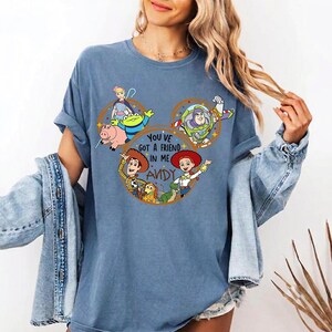 May include: A blue t-shirt featuring a graphic of Toy Story characters arranged in a Mickey Mouse ear shape. The design includes Woody, Buzz Lightyear, Jessie, Bo Peep, Rex, Slinky Dog, and Hamm, with the text "YOU'VE GOT A FRIEND IN ME ANDY".