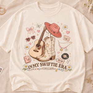 Puede incluir: Una camiseta de color crema con un estampado gr&aacute;fico de una guitarra ac&uacute;stica, una bota vaquera, un sombrero rosa, gafas de sol en forma de coraz&oacute;n, un casete y flores. El texto "IN MY SWIFTIE ERA" y "TAYLOR" aparece impreso en letras marrones.