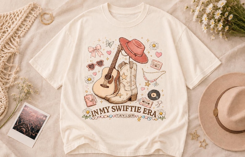 Puede incluir: Una camiseta de color crema con un estampado gr&aacute;fico de una guitarra ac&uacute;stica, una bota vaquera, un sombrero rosa, gafas de sol en forma de coraz&oacute;n, un casete y flores. El texto "IN MY SWIFTIE ERA" y "TAYLOR" aparece impreso en letras marrones.