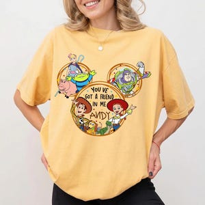 May include: A mustard yellow short-sleeve t-shirt featuring a graphic print of Toy Story characters arranged in circles. The text reads YOU VE GOT A FRIEND IN ME ANDY. Characters include Woody, Buzz Lightyear, Jessie, Bo Peep, and others.