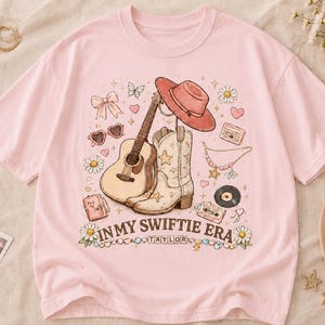 Puede incluir: Una camiseta de manga corta de color rosa claro con un estampado gr&aacute;fico de una guitarra ac&uacute;stica, una bota vaquera, un sombrero vaquero, un casete, un disco de vinilo, corazones, flores y un lazo. El texto dice IN MY SWIFTIE ERA y TAYLOR.