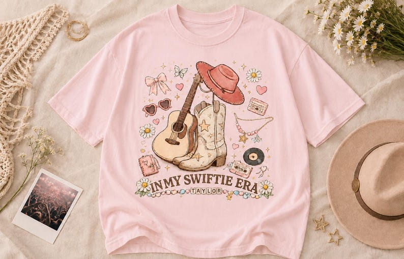 Puede incluir: Una camiseta de manga corta de color rosa claro con un estampado gr&aacute;fico de una guitarra ac&uacute;stica, una bota vaquera, un sombrero vaquero, un casete, un disco de vinilo, corazones, flores y un lazo. El texto dice IN MY SWIFTIE ERA y TAYLOR.