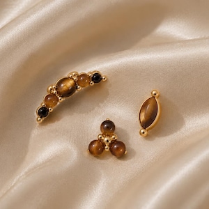 May include: Three gold-toned earrings featuring brown tiger's eye and black stones. One earring is a curved bar, one is a triangular cluster, and one is a marquise-shaped stud. Each piece is crafted with polished metal settings and smooth cabochon stones.