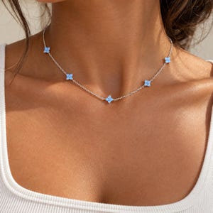 May include: A silver chain necklace featuring five evenly spaced, four-petaled flower charms. Each flower charm is set with light blue stones that have a subtle iridescent sheen. The necklace rests against the collarbone area.