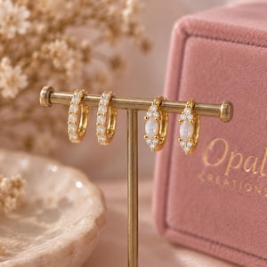 May include: Two pairs of gold hoop earrings displayed on a metal stand. The first pair features a row of small round opals, while the second pair showcases a central oval opal accented by tiny clear stones. The jewelry is presented near a pink box.