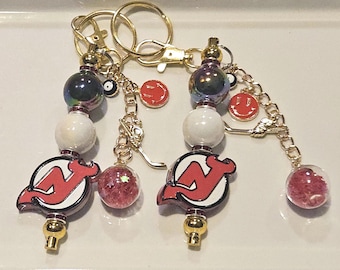 May include: Two gold-toned keychains featuring a New Jersey Devils logo charm, iridescent dark beads, white spherical beads, and small dangling charms including a red smiley face, a miniature hockey stick, and a clear orb filled with pink glitter.
