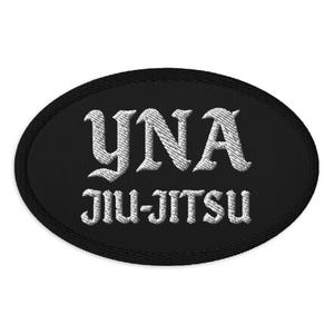 May include: Black oval patch with white embroidered text. The text reads "YNA" above "JIU-JITSU" in a stylized font. The patch has a black border and is likely for clothing or gear.