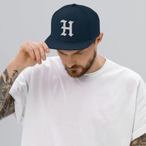 May include: A navy blue baseball cap with a flat brim. The cap features a white, stylized letter "H" embroidered on the front. The hat is worn by a person wearing a white t-shirt.
