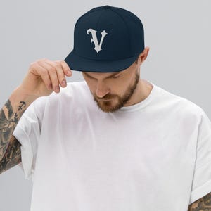 May include: A navy blue baseball cap with a white, stylized letter "V" embroidered on the front. The cap has a flat brim and a structured fit. The person in the image is wearing a white t-shirt.
