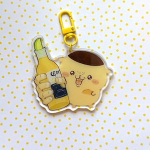 May include: A yellow acrylic keyring featuring a cartoon pudding character holding a Corona beer bottle with a lime wedge. The keyring has a yellow clasp and ring. The background is white with yellow polka dots.