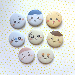 Peut inclure: Huit boutons ronds et colorés avec des visages de dessins animés. Chaque bouton présente une expression et un design différents, notamment des cheveux bleus, une étoile et un visage de koala. Les boutons sont disposés sur une surface blanche à pois jaunes.