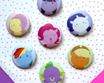 May include: A collection of seven round, colorful buttons featuring cartoon pony characters. Each button has a different character design with various colors, including purple, pink, blue, yellow, and green. The buttons are arranged on a polka dot background.