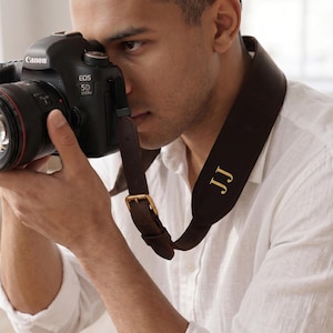 May include: A dark brown leather camera strap with a gold-toned metal buckle and the gold-colored initials JJ embossed on the surface. The strap is attached to a black Canon EOS 5D camera body with a lens, featuring a red ring around the lens barrel.