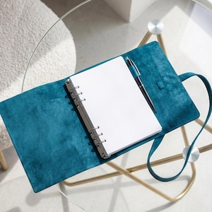 May include: A teal suede ring-bound planner with a matching strap closure and a silver metal six-ring mechanism. The planner is open to reveal blank white pages and includes a black pen held in a dedicated loop on the interior cover.