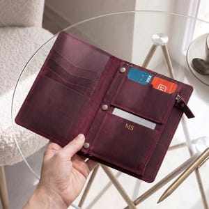 Puede incluir: Cartera de cuero burdeos abierta, mostrando ranuras para tarjetas, un bolsillo con cremallera y un bolsillo pequeño con una tarjeta. Las iniciales "MS" están grabadas en la parte delantera. Se ve una Visa y una tarjeta de crédito.