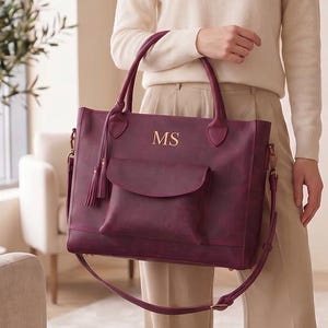 Op de afbeelding: Een diepbordeauxrode tote bag met een voorvak met klep, twee handvatten en een afneembare schouderriem. De tas is gepersonaliseerd met de goudkleurige initialen MS op de voorzijde en is voorzien van een decoratief kwastje aan een handvat.