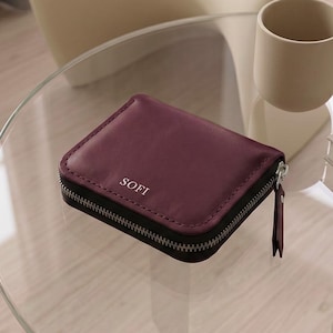 May include: A burgundy, square wallet with the word "SOFI" printed on the front. The wallet has a black zipper and a small pull tab. It sits on a glass table next to a beige cup.