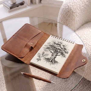 May include: A brown leather notebook cover with a snap closure, containing a spiral-bound sketchbook with a black and white ink illustration of an oak tree and acorns on the cover page, accompanied by a matching brown wooden ballpoint pen.