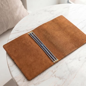 May include: A rectangular brown leather notebook cover featuring a soft, suede-like texture. The interior includes three light blue elastic cords secured by gold-toned metal eyelets, designed to hold multiple notebook inserts in place.