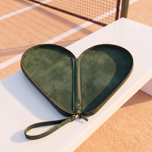 May include: A heart-shaped dark green case with a smooth interior lining and a matching wrist strap. The case features a central zipper closure and a structured frame, designed to open fully to reveal two symmetrical compartments for storage.