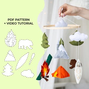 May include: A handmade felt baby mobile featuring camping-themed elements. The mobile includes a campfire, tent, bear, kayak, mountains, trees, and clouds. The image also shows a pattern with the text "PDF PATTERN + VIDEO TUTORIAL".