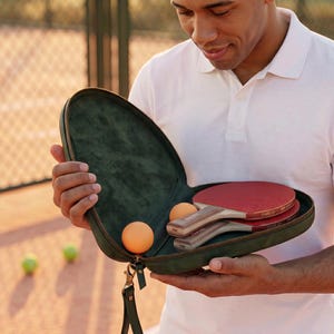 May include: A dark green, oval-shaped zippered carrying case containing two table tennis paddles with red rubber surfaces and wooden handles, along with two orange table tennis balls nestled inside the velvet-lined interior compartment.