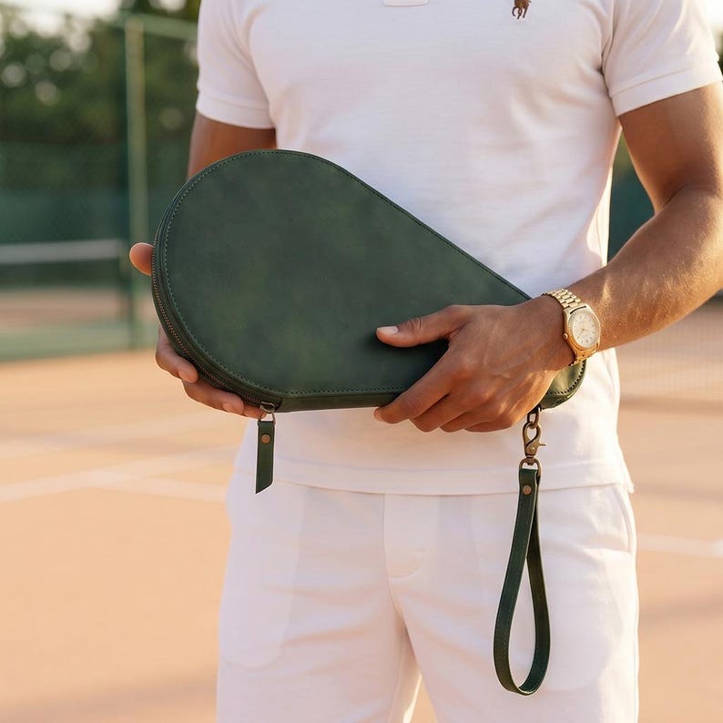May include: A dark green, teardrop-shaped leather case designed for a pickleball or tennis paddle. The case features a secure zipper closure, a matching leather wrist strap with a metal clasp, and visible stitching along the curved edges.
