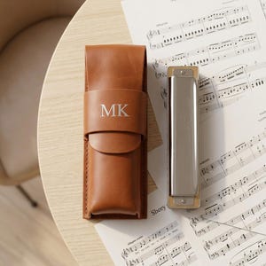 Puede incluir: Un estuche de cuero marrón para armónica con las iniciales "MK" grabadas en la parte delantera. Junto al estuche, una armónica plateada con detalles dorados, sobre partituras. El estuche está diseñado para guardar la armónica.