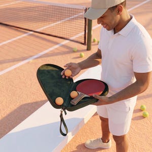May include: A dark green zippered carrying case containing a wooden paddle with a red rubber surface and three orange balls. The case features a matching green wrist strap and a soft interior lining, designed to hold the paddle and balls securely.