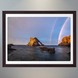 Peut inclure: Tirage encadré d'un paysage côtier avec un double arc-en-ciel au-dessus de la mer. L'image présente une formation rocheuse naturelle avec une arche, éclairée par le soleil couchant. Le ciel passe du bleu au rose.