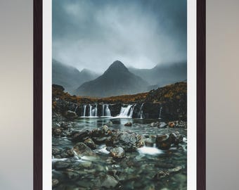 May include: Framed landscape print depicting a mountain range under a cloudy sky. A waterfall flows into a rocky stream in the foreground. The print is in a dark brown frame with a white border, creating a striking contrast.