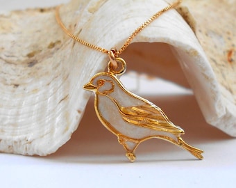May include: A gold necklace with a white enamel bird pendant. The necklace is displayed on a white seashell.