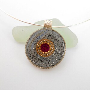 May include: A silver necklace with a round pendant. The pendant is made of gray resin with a sparkly finish and a red rhinestone in the center surrounded by gold rhinestones.