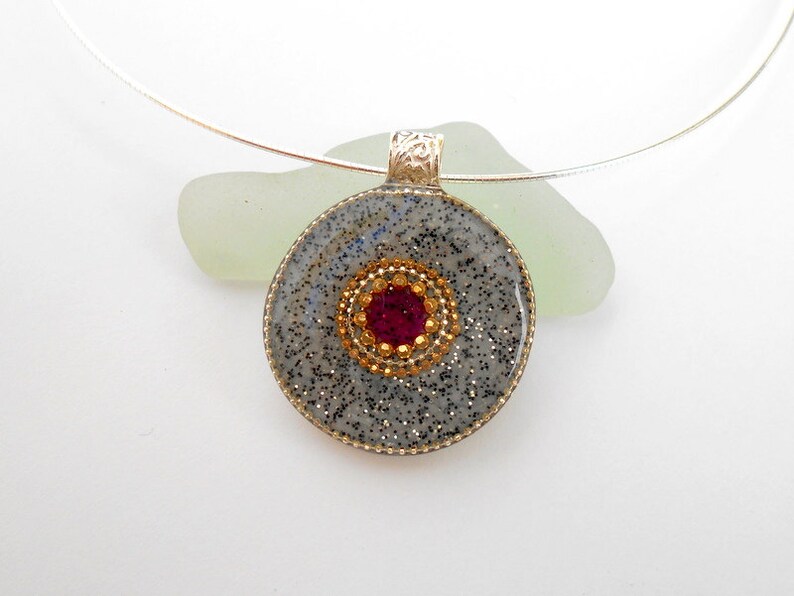 May include: A silver necklace with a round pendant. The pendant is made of gray resin with a sparkly finish and a red rhinestone in the center surrounded by gold rhinestones.