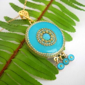 Puede incluir: Un collar con un colgante turquesa, una cadena de oro y un charm redondo turquesa con un centro de oro y &oacute;palo. El charm tiene un borde de cuentas y tres charms turquesa m&aacute;s peque&ntilde;os que cuelgan.