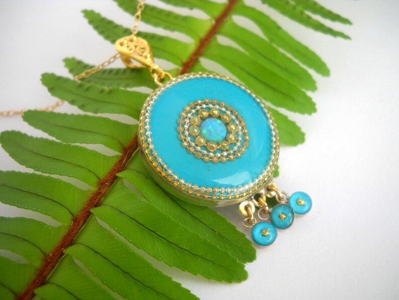 Puede incluir: Un collar con un colgante turquesa, una cadena de oro y un charm redondo turquesa con un centro de oro y &oacute;palo. El charm tiene un borde de cuentas y tres charms turquesa m&aacute;s peque&ntilde;os que cuelgan.