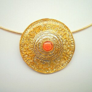 Puede incluir: Un collar de oro con un colgante redondo, de dise&ntilde;o ornamentado, que presenta una peque&ntilde;a piedra preciosa de color rojo anaranjado en el centro.