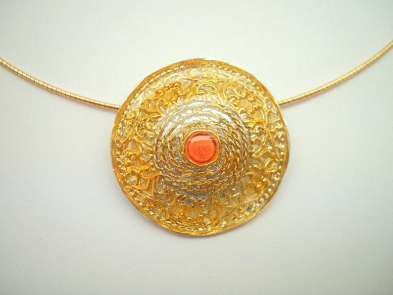 Puede incluir: Un collar de oro con un colgante redondo, de dise&ntilde;o ornamentado, que presenta una peque&ntilde;a piedra preciosa de color rojo anaranjado en el centro.