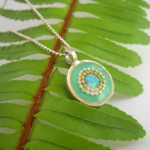 May include: A silver chain necklace with a round pendant featuring a turquoise blue resin background with a gold and silver beaded circle surrounding a light blue opal.