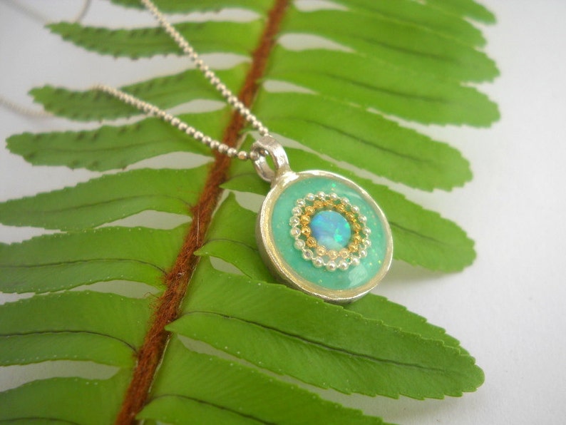May include: A silver chain necklace with a round pendant featuring a turquoise blue resin background with a gold and silver beaded circle surrounding a light blue opal.