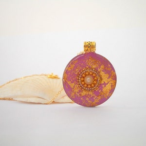 May include: A pink and gold round pendant necklace with a pearl in the center. The pendant is made of resin and has a gold leaf design.