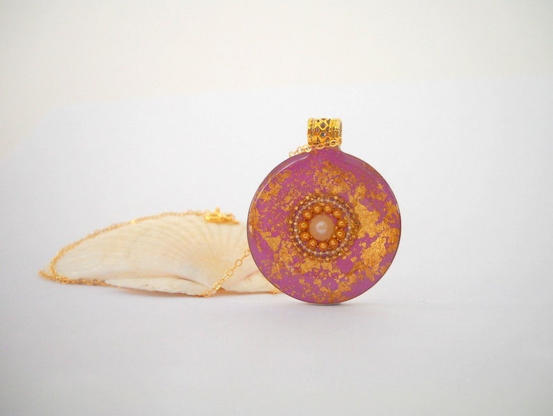 May include: A pink and gold round pendant necklace with a pearl in the center. The pendant is made of resin and has a gold leaf design.