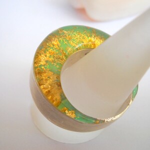 May include: A green and gold resin ring with a unique, marbled design. The ring is displayed on a white ring stand.