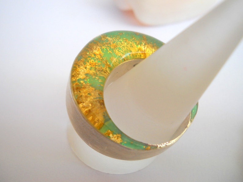 May include: A green and gold resin ring with a unique, marbled design. The ring is displayed on a white ring stand.