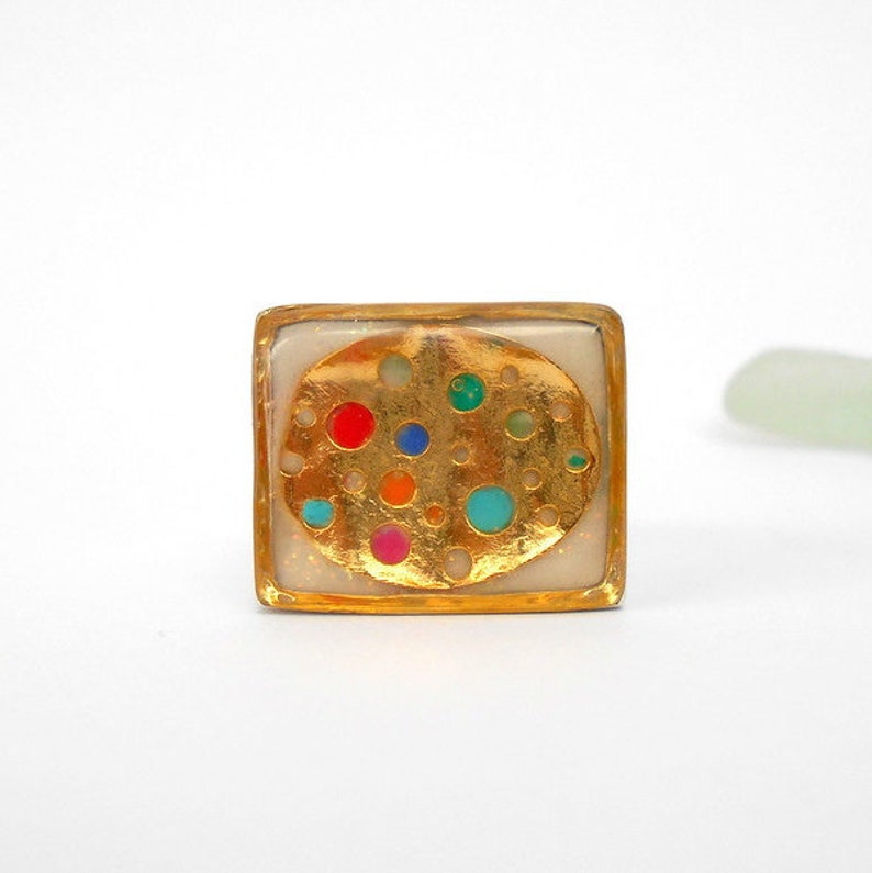 May include: Square gold ring with a white background and a gold circle with colorful dots inside.