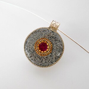 May include: A silver pendant with a round, gray, glitter-covered resin charm. The charm has a gold-colored circle with a red rhinestone in the center.