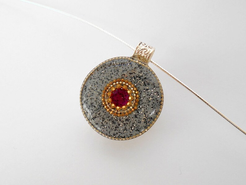 May include: A silver pendant with a round, gray, glitter-covered resin charm. The charm has a gold-colored circle with a red rhinestone in the center.